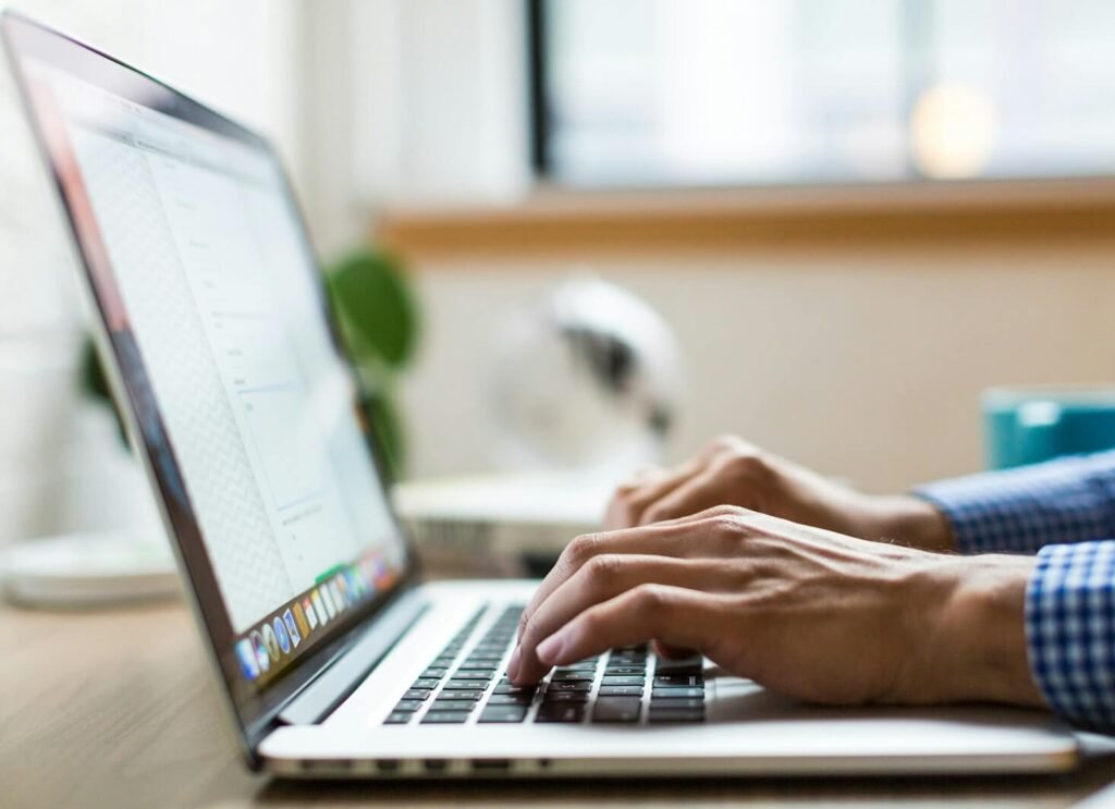 Mains d'un homme tapant sur le clavier d'un MacBook dans un environnement de bureau lumineux, avec une plante verte visible en arrière-plan, illustrant le travail de bureau moderne