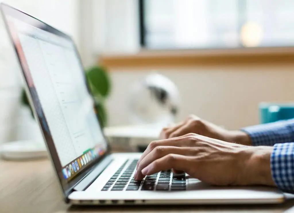 Mains d'un homme tapant sur le clavier d'un MacBook dans un environnement de bureau lumineux, avec une plante verte visible en arrière-plan, illustrant le travail de bureau moderne