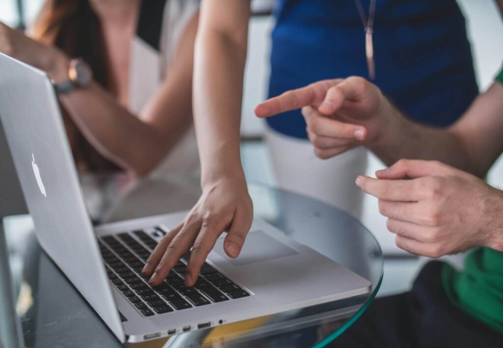 Scène de collaboration en entreprise où plusieurs personnes discutent autour d'un ordinateur portable, avec des gestes explicatifs, dans un environnement de travail moderne et lumineux
