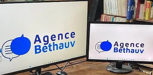 Deux écrans d'ordinateur côte à côte affichant le logo "Agence Bethauv" avec un symbole de bulle de dialogue bleue, dans un bureau avec des livres en arrière-plan.